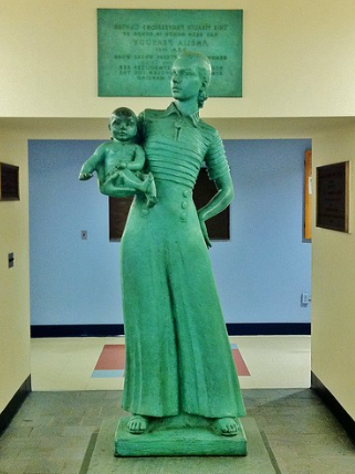 一张绿色金属雕像的彩色照片. 雕像是一个女人用一只胳膊抱着一个婴儿的形象. 她穿着一条波浪形的长裤，上面有六颗纽扣，还有一条条纹, 件衬衫, 条纹是有纹理的. 她在一栋学校大楼里，周围都是瓷砖地板和牌匾, 包括她的头顶和背后，观众可以看到艺术家的名字, 阿米莉亚皮博迪, 但它太模糊了，看不清其他的字.